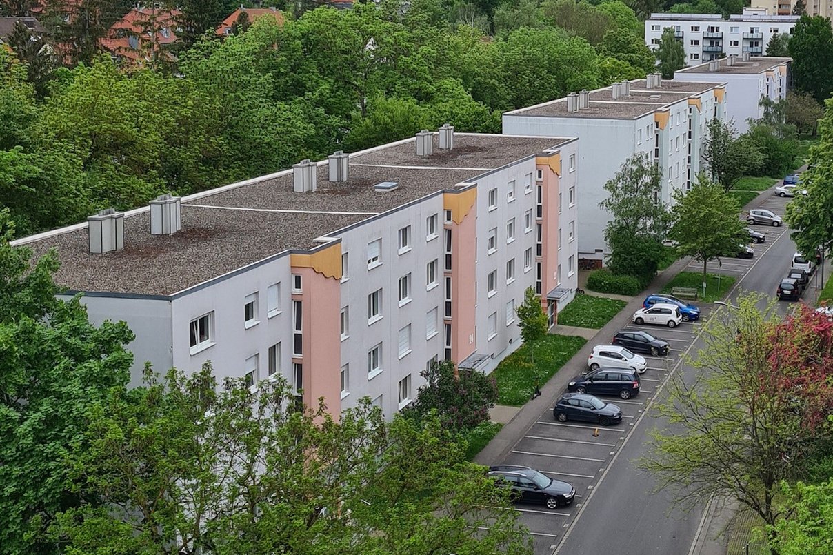 Vogelperspektive auf eine Wohnsiedlung mit mehreren weiß-grauen Mehrfamilienhäusern, begrünten Außenanlagen und einer Straße mit parkenden Autos. Im Hintergrund sind weitere Wohnhäuser und dichtes, grünes Baumwerk zu sehen.