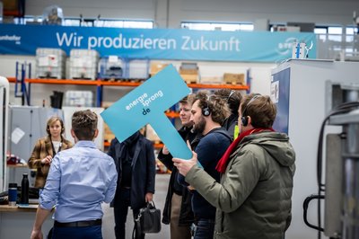 Mehrere Personen stehen in einem Forschungsraum. Einige Personen tragen Kopfhörer auf dem Kopf. Eine Person hält blaues Schild in der Hand mit Aufschrift: energiesprong.de. Im Hintergrund ein Banner mit Text: Wir produzieren Zukunft.