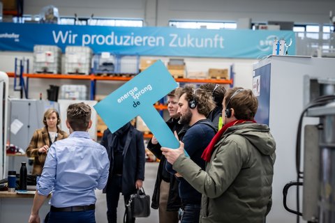Mehrere Personen stehen in einem Forschungsraum. Einige Personen tragen Kopfhörer auf dem Kopf. Eine Person hält blaues Schild in der Hand mit Aufschrift: energiesprong.de. Im Hintergrund ein Banner mit Text: Wir produzieren Zukunft.