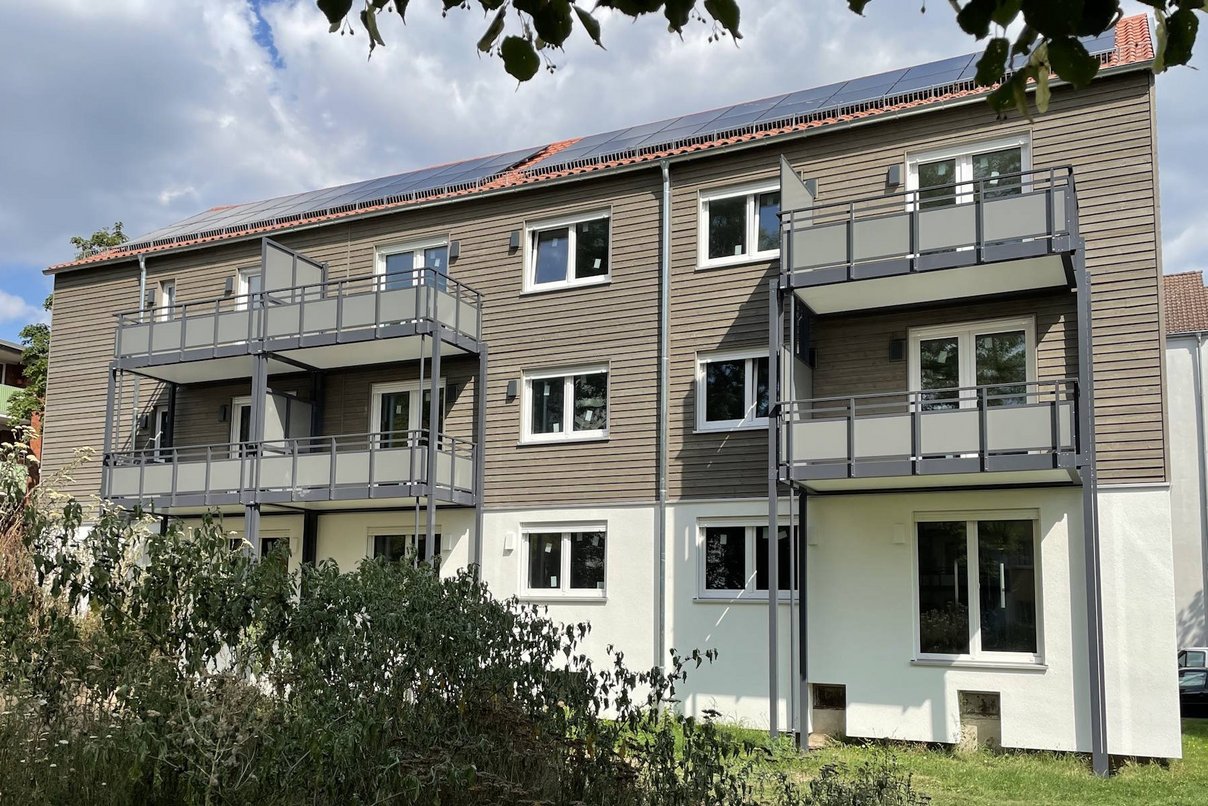 Mehrfamiienhaus dreigeschossig mit Balkonen, davor Grünanlagen, im Hintergrund bewölkter Himmel