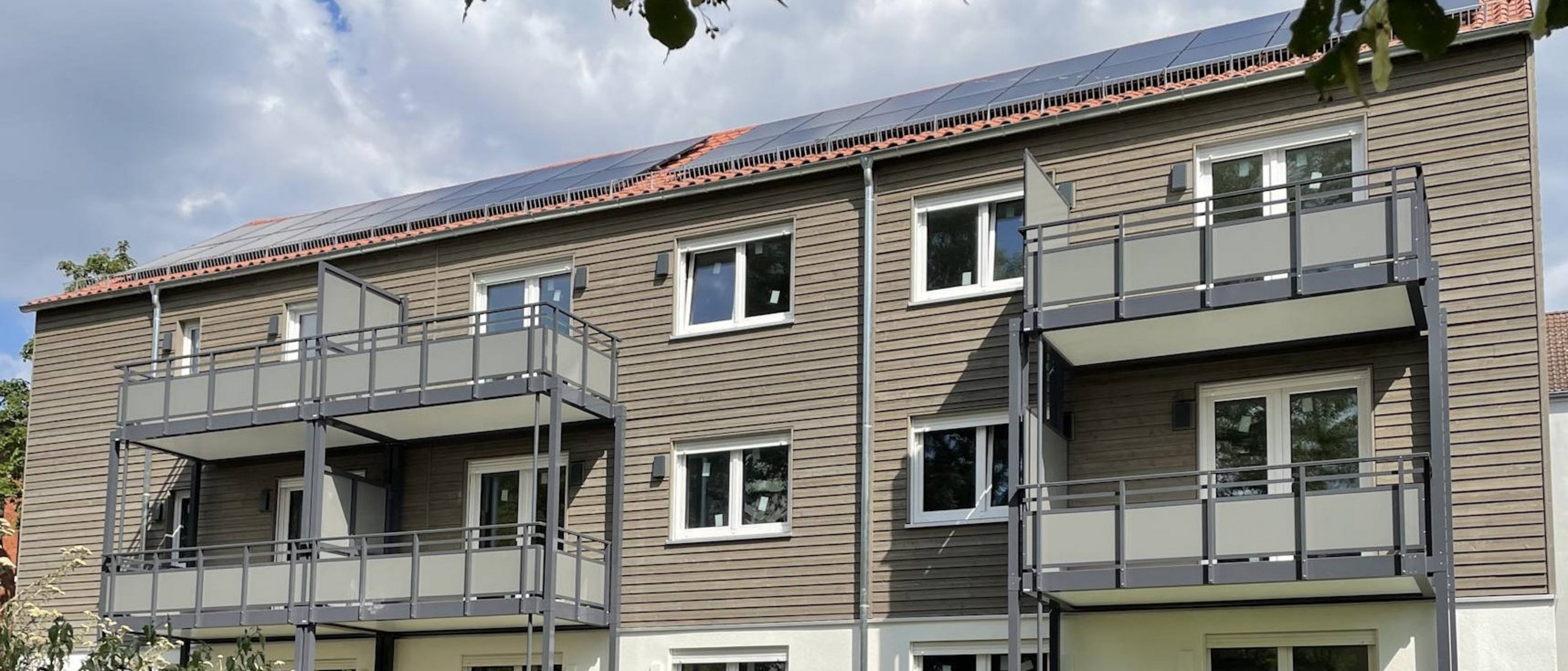 Mehrfamiienhaus dreigeschossig mit Balkonen, davor Grünanlagen, im Hintergrund bewölkter Himmel