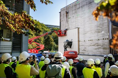 Bild zeigt Menschengruppe vor Gebäudefassade an der im Moment ein Kran ein neues Fassadenelement montiert