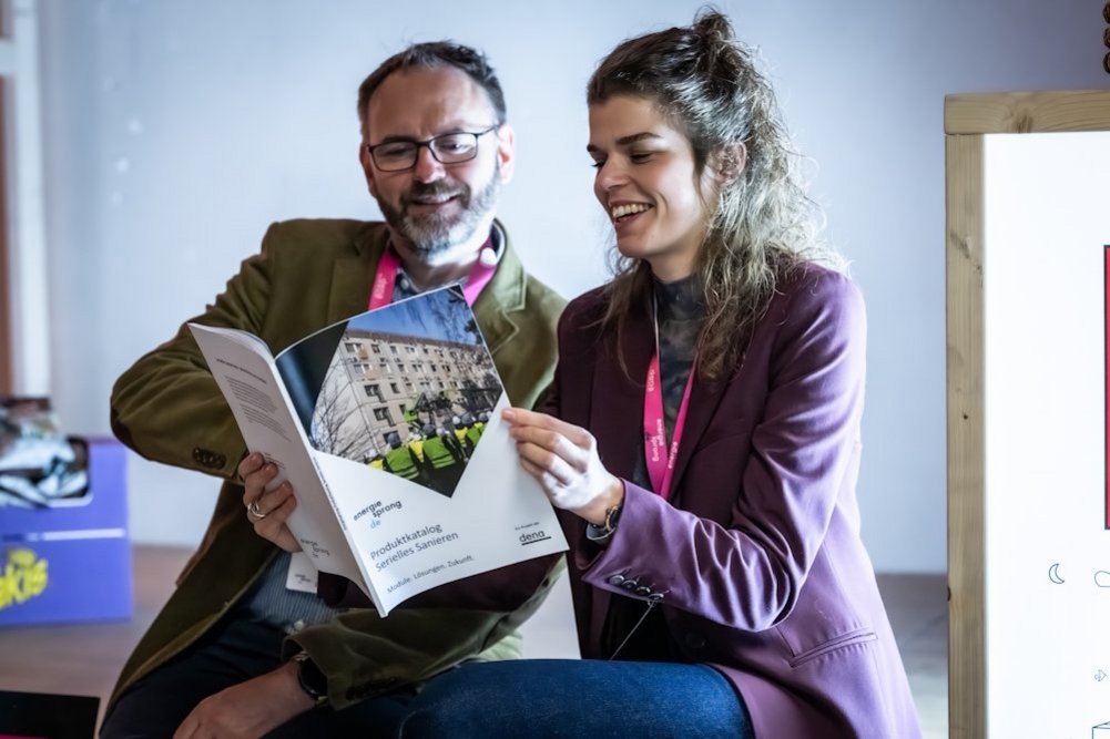 Zwei Personen sitzen nebeneinander und schauen gemeinsam in eine Broschüre mit der Aufschrift "Produktkatalog Serielles Sanieren".