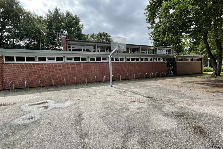 Außenansicht einer Schulturnhalle, davor eine betonierte Fläche mit Basketballkorb