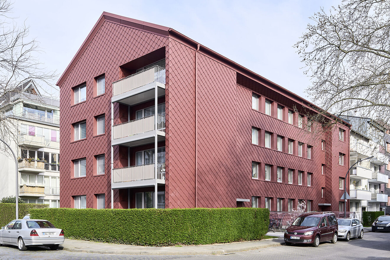 Saniertes Mehrfamilienhaus mit Fassade aus roten Alu-Rauten, davor Straße mit PKWs