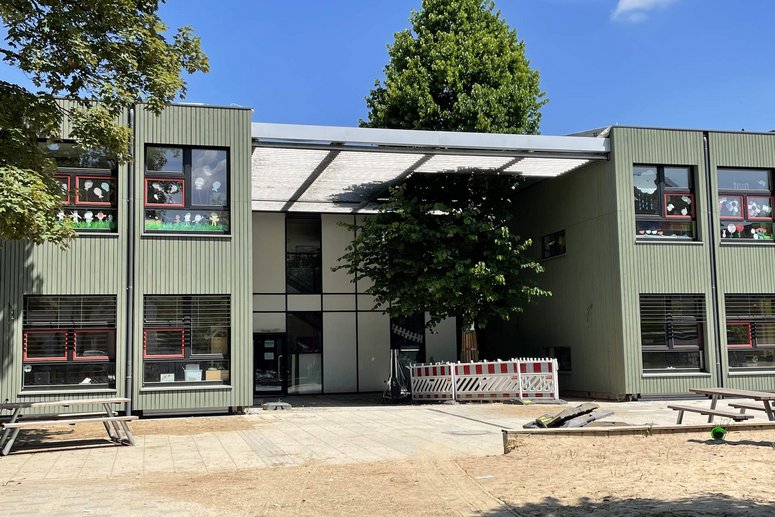 Kita-Gebäude zweigeschossig mit hellgrüner Holzfassade, überdachter Eingangsbereich mit großem Laubbaum