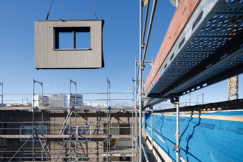 Vorgefertigtes Fassadenelement mit Fenster schwebt an Seilen über Gebäude mit Baugerüst, im Hintergrund blauer Himmel