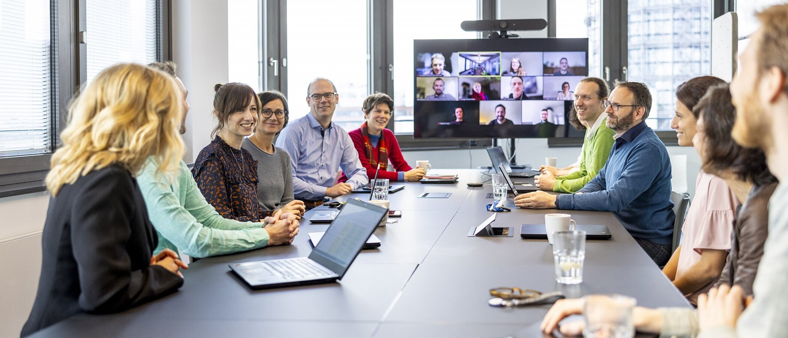 Mehrere Personen am Besprechungstisch, auf dem Tisch sind Notebooks, im Hintergrund ein großer Bildschirm