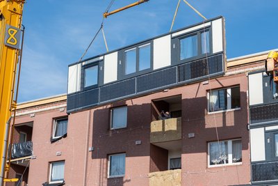 Vorgefertigtes Fassdaenlement schwebt an einem Kran vor der Fassade eines Mehrfamilienhauses.