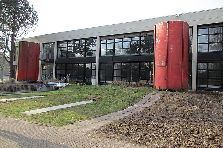 Schulgebäude zweigeschossig mit Flachdach unsaniert, davor stehen zwei große rote Tanks.