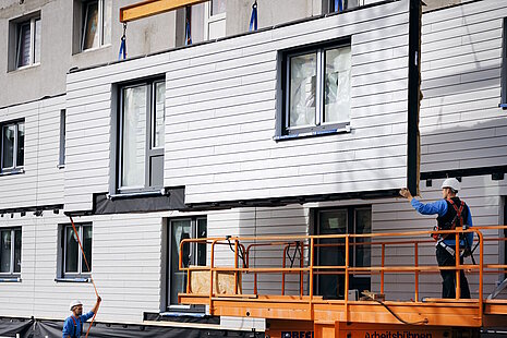 Mehrfamilienhaus während Sanierung, schwebendes, vorgefertigtes Fassadenlement, Bauarbeiter