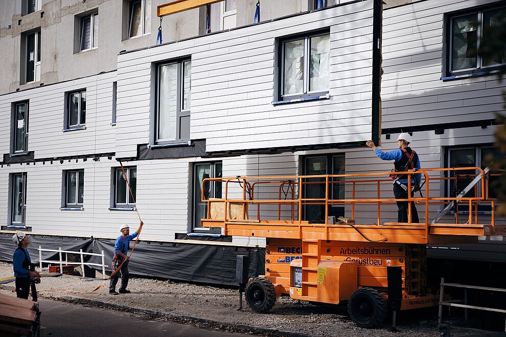 Mehrfamilienhaus während Sanierung, schwebendes, vorgefertigtes Fassadenlement, Bauarbeiter