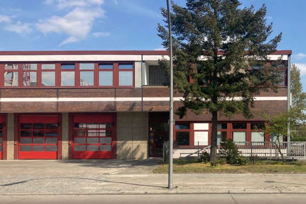 Feuerwache , davor ein Baum und eine Straßenlaterne, blauer Himmel mit einzelnen Wolken