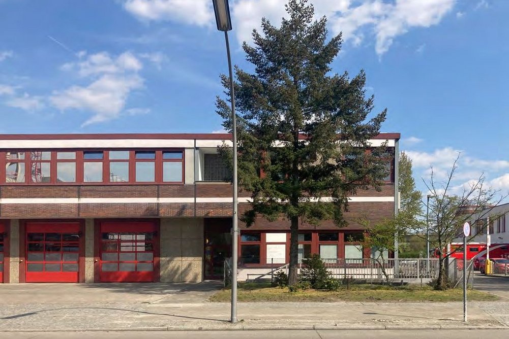 Feuerwache , davor ein Baum und eine Straßenlaterne, blauer Himmel mit einzelnen Wolken