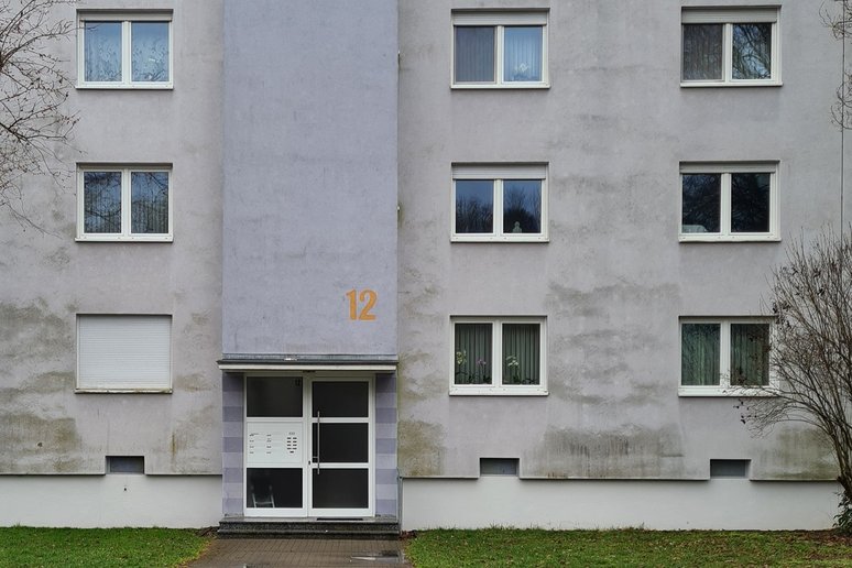 Frontansicht eines dreigeschossigen Mehrfamilienhauses mit hellgrauer, teilweise verwitterter Fassade. In der Mitte befindet sich der Hauseingang mit der orangefarbenen Hausnummer 12. Mehrere weiße Fenster und Rollläden sind regelmäßig über die Fassade verteilt; vor dem Gebäude liegt eine schmale Rasenfläche.