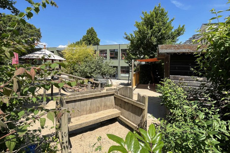 Kita-Spielplatz mit Laubbäumen und Sträuchern, im Hintergrund Kita-Gebäuse zweigeschossig, blauer wolkenloser Himmel