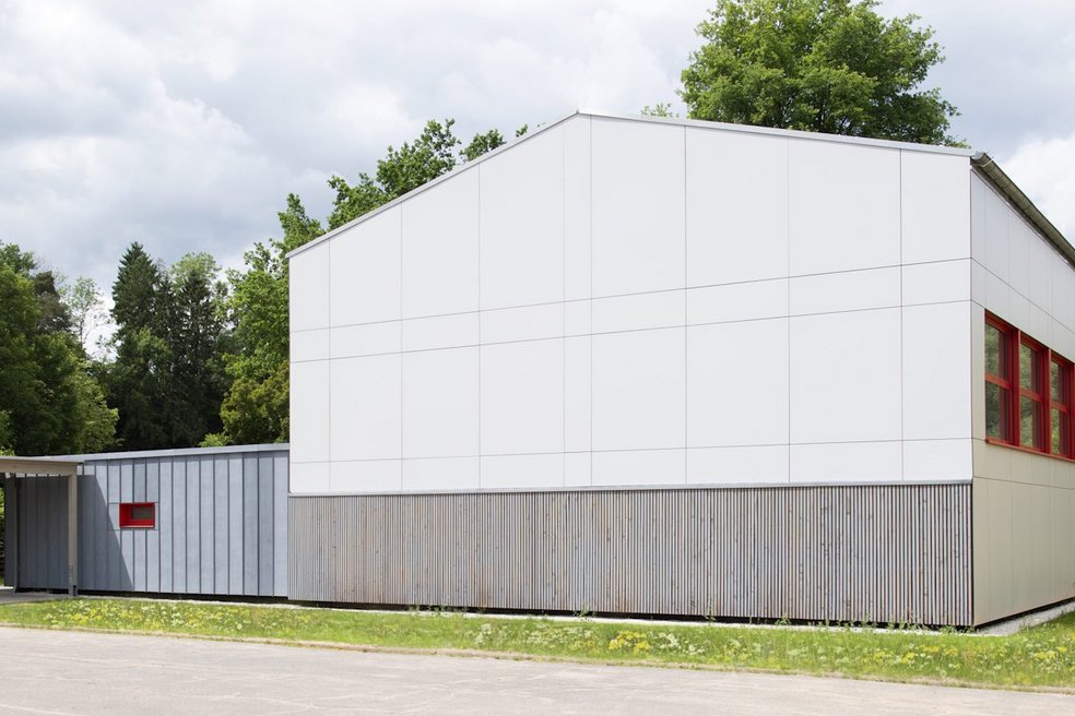 Außenansicht neu sanierte Turnhalle umgeben von Rasenflächen und Laubbäumen, davor eine große betonierte Fläche
