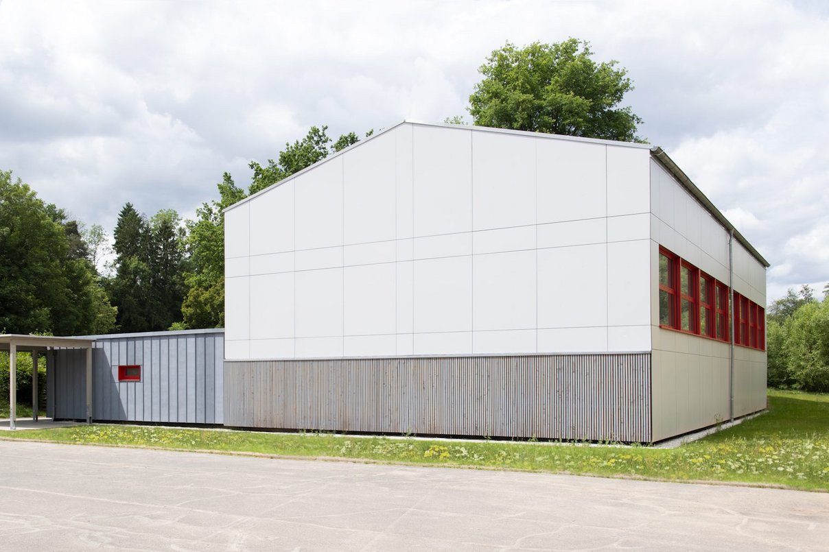 Außenansicht neu sanierte Turnhalle umgeben von Rasenflächen und Laubbäumen, davor eine große betonierte Fläche