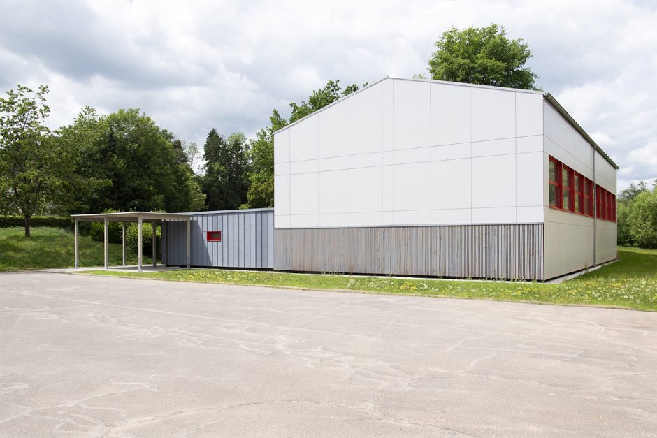 Außenansicht neu sanierte Turnhalle umgeben von Rasenflächen und Laubbäumen, davor eine große betonierte Fläche