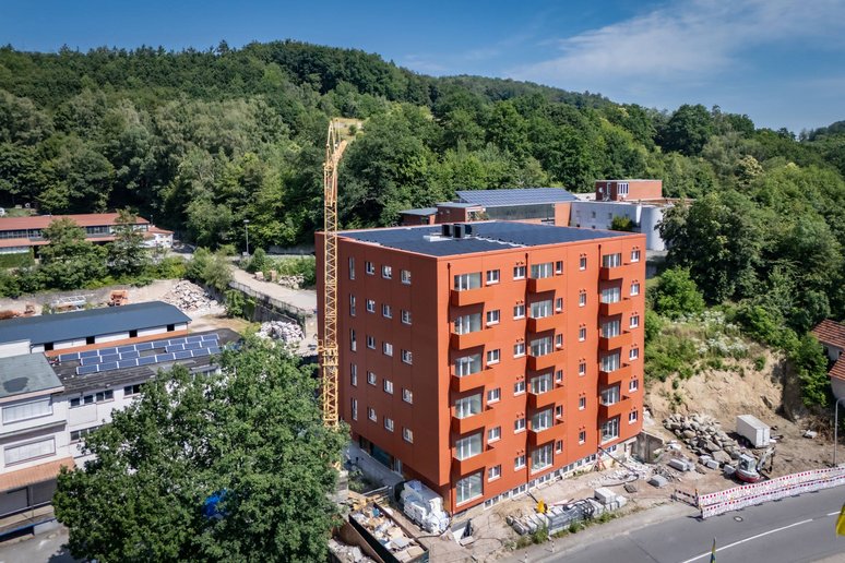 Blick aus der Luft auf ein Gebäude sechsgeschossig während Sanierung, davor eine Straße, im Hintergrund Hügellandschaft mit Laubbäumen, seitlich vom Gebäude weitere Gebäude