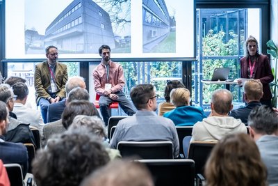 Personen sitzen auf Stühlen in einem Veranstaltungsraum und schauen zur Bühne.