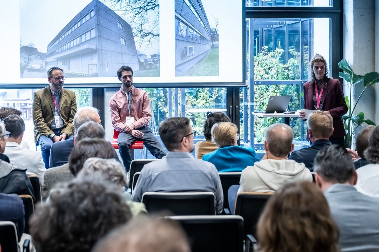 Personen sitzen auf Stühlen in einem Veranstaltungsraum und schauen zur Bühne.