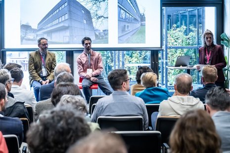 Personen sitzen auf Stühlen in einem Veranstaltungsraum und schauen zur Bühne.