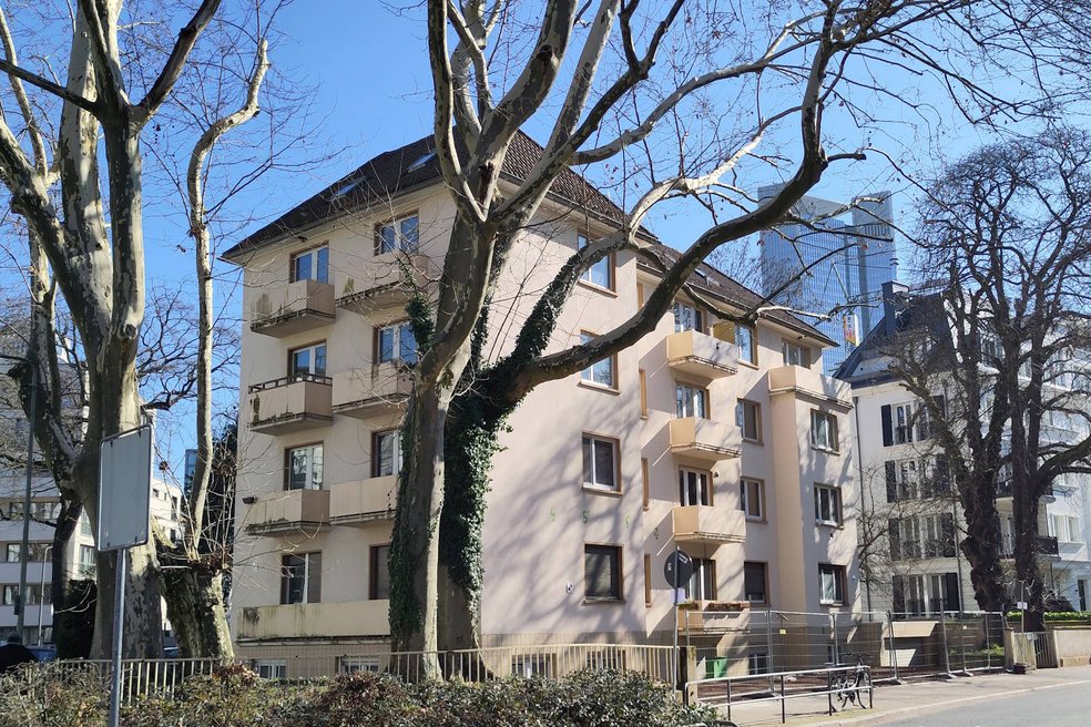 Verschieben Sie den Pfeil für den Vorher/Nachher-Vergleich Mehrfamilienhaus viergeschossig mit kleinen Balkonen, unsaniert. Davor eine Straße mit Bäumen ohne Blätter.