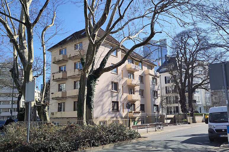 Mehrfamilienhaus viergeschossig mit kleinen Balkonen, unsaniert. Davor eine Straße mit Bäumen ohne Blätter.