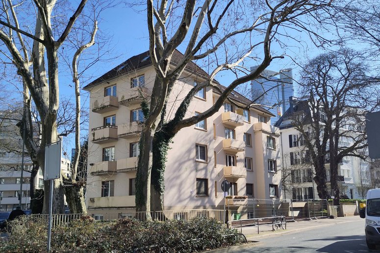 Mehrfamilienhaus viergeschossig mit kleinen Balkonen, unsaniert. Davor eine Straße mit Bäumen ohne Blätter.
