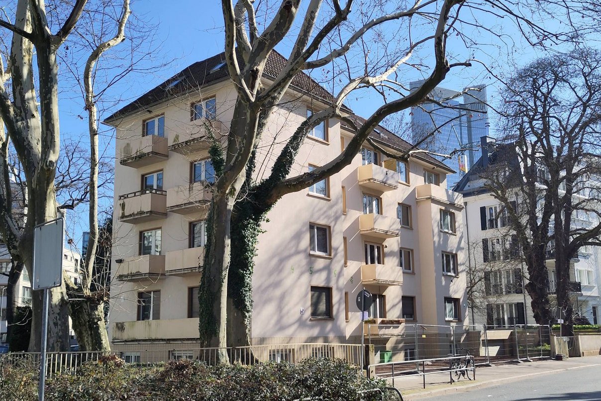 Mehrfamilienhaus viergeschossig mit kleinen Balkonen, unsaniert. Davor eine Straße mit Bäumen ohne Blätter.