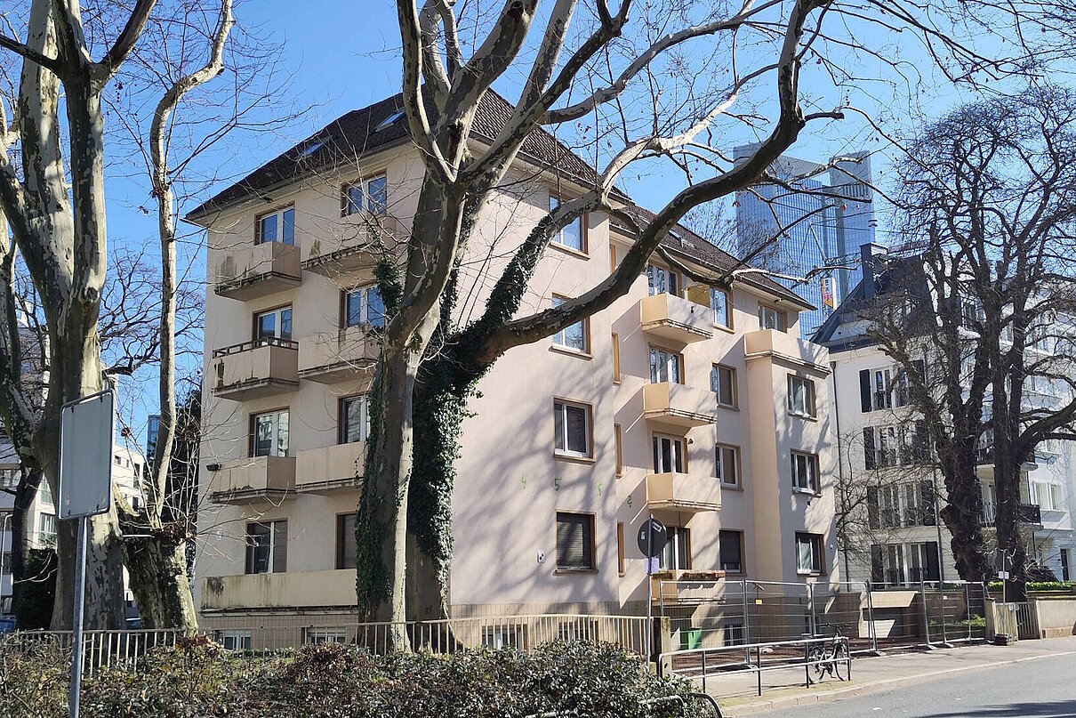 Mehrfamilienhaus viergeschossig mit kleinen Balkonen, unsaniert. Davor eine Straße mit Bäumen ohne Blätter.