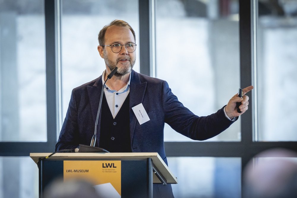 Marc Becker Person in Business-Kleidung steht am Pult und redet.