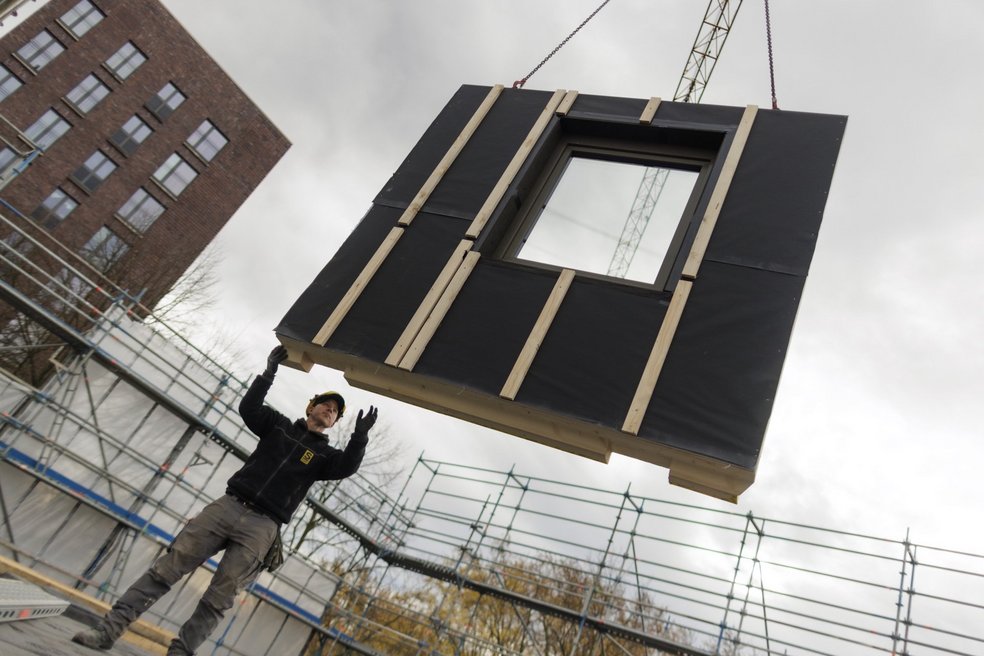 Vorgefertigtes Hausfassadenelement, welches an einem Kran schwebt und von einem Bauarbeiter mit den Händen navigiert wird.