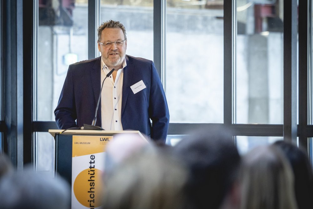 Alexander Fuchs Person in Business-Kleidung steht hinter Rednerpult, davor sitzt Publikum.