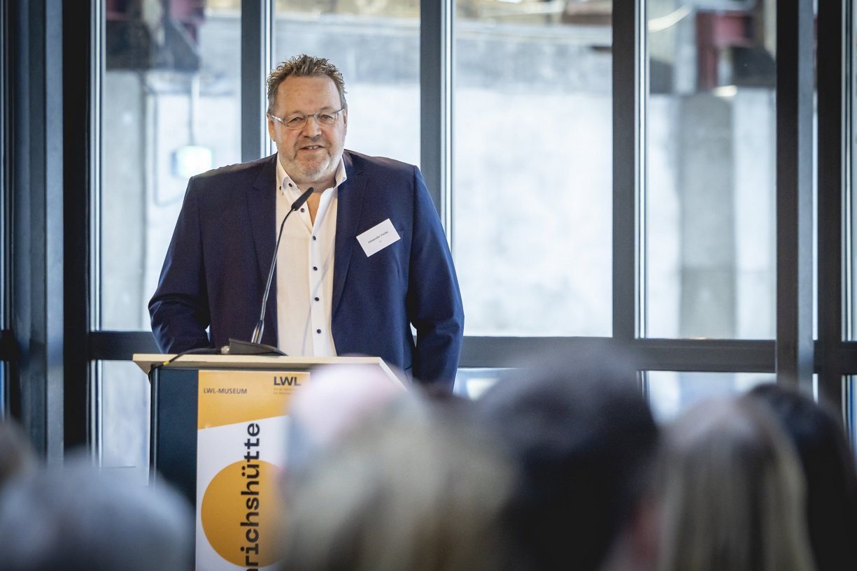 Alexander Fuchs Person in Business-Kleidung steht hinter Rednerpult, davor sitzt Publikum.
