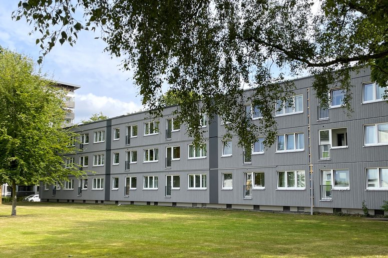 Modernes mehrstöckiges Gebäude mit grauer Fassade und vielen Fenstern, umgeben von einem gepflegten Rasen und Bäumen unter einem leicht bewölkten Himmel.