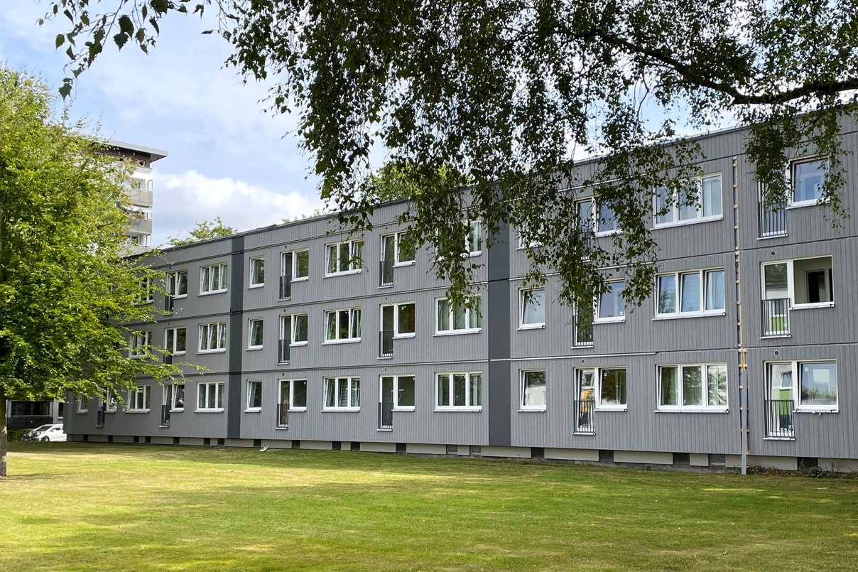 Modernes mehrstöckiges Gebäude mit grauer Fassade und vielen Fenstern, umgeben von einem gepflegten Rasen und Bäumen unter einem leicht bewölkten Himmel.