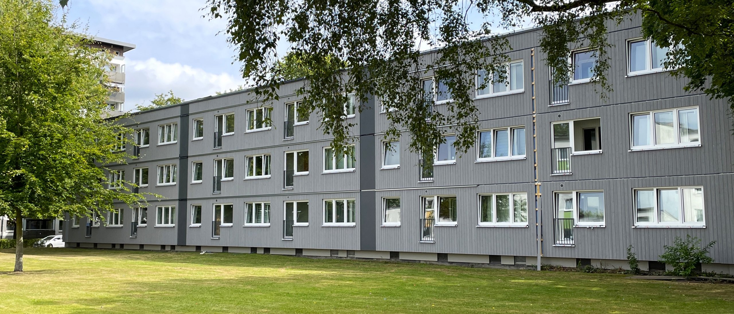 Modernes mehrstöckiges Gebäude mit grauer Fassade und vielen Fenstern, umgeben von einem gepflegten Rasen und Bäumen unter einem leicht bewölkten Himmel.