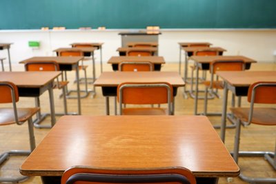 Klassenraum ohne Kinder, im vordegrund Tische mit Stühlen aus Holz, im Hintergrund breite Tafelfläche (grün)