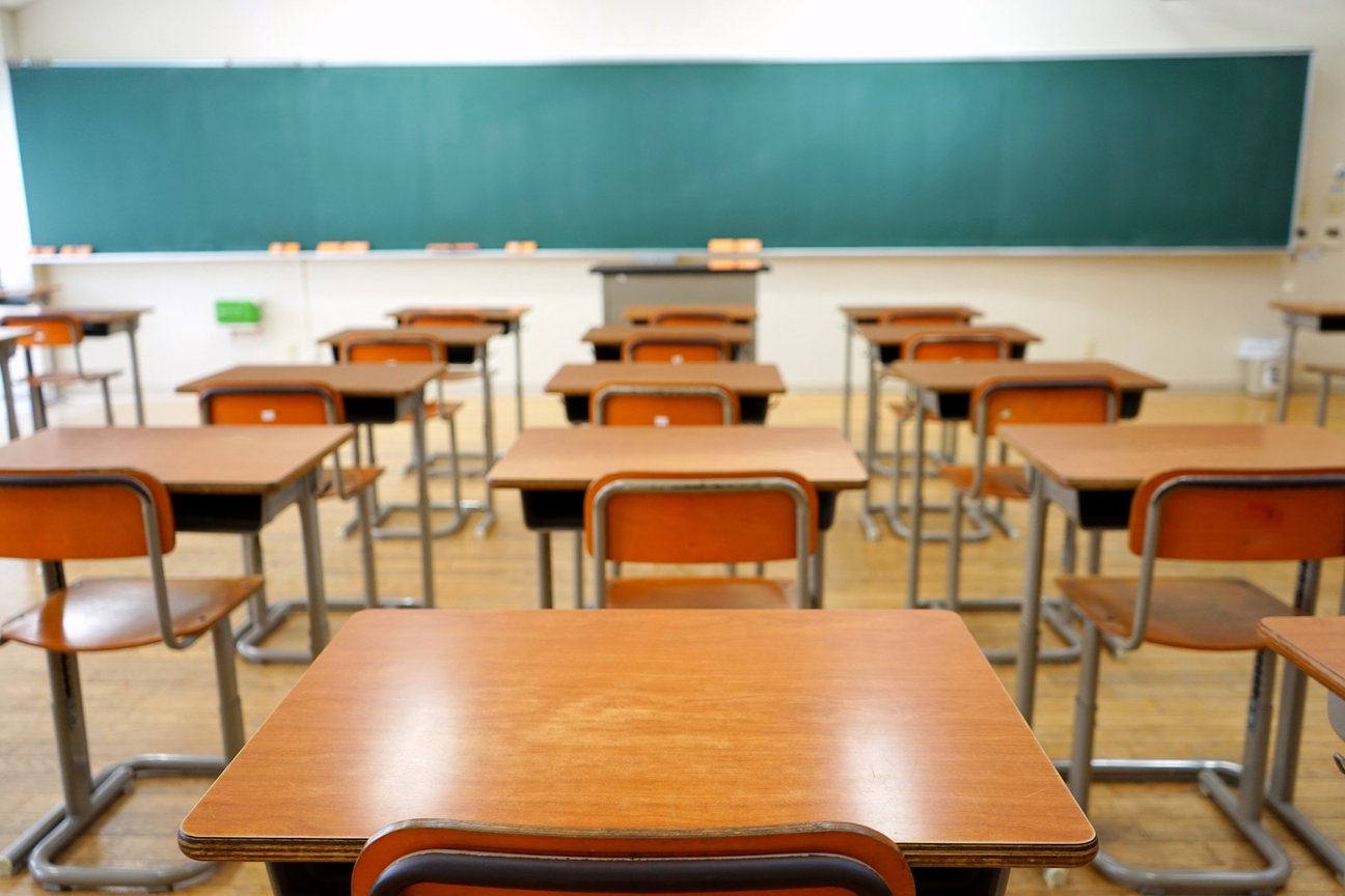 Klassenraum ohne Kinder, im vordegrund Tische mit Stühlen aus Holz, im Hintergrund breite Tafelfläche (grün)