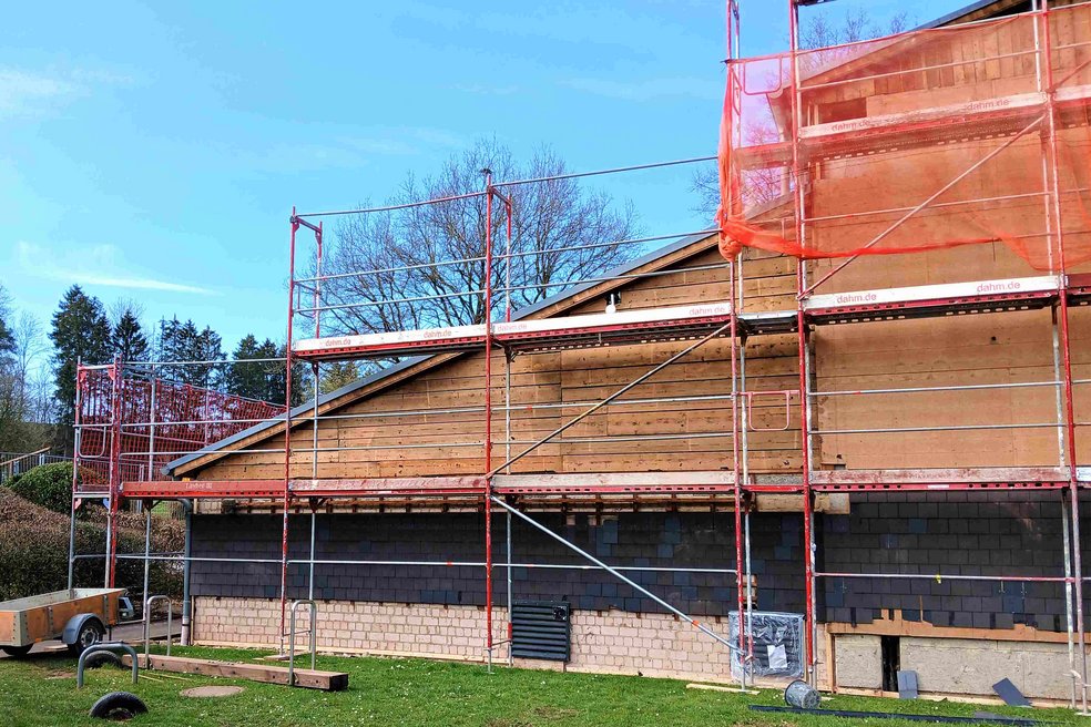 Außenansicht Turnhalle mit Schrägdach während Sanierung, davor Baugerüst und Grünflächen