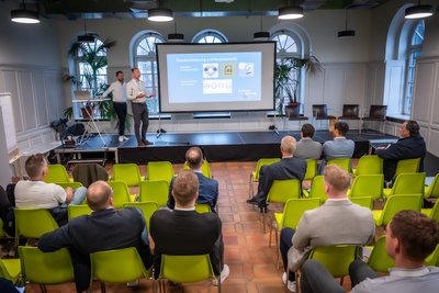 Personen sitzen in einem Raum auf leuchtend grünen Stühlen und blicken zu den Referenten auf der Bühne.