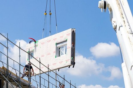 Vorgefertigtes Hausfassadenelement schwebt an einem Kran, Haus mit Baugerüst, zwei Bauarbeiter