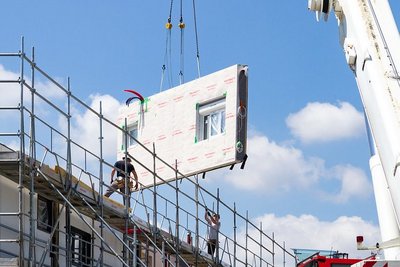 Vorgefertigtes Hausfassadenelement schwebt an einem Kran, Haus mit Baugerüst, zwei Bauarbeiter