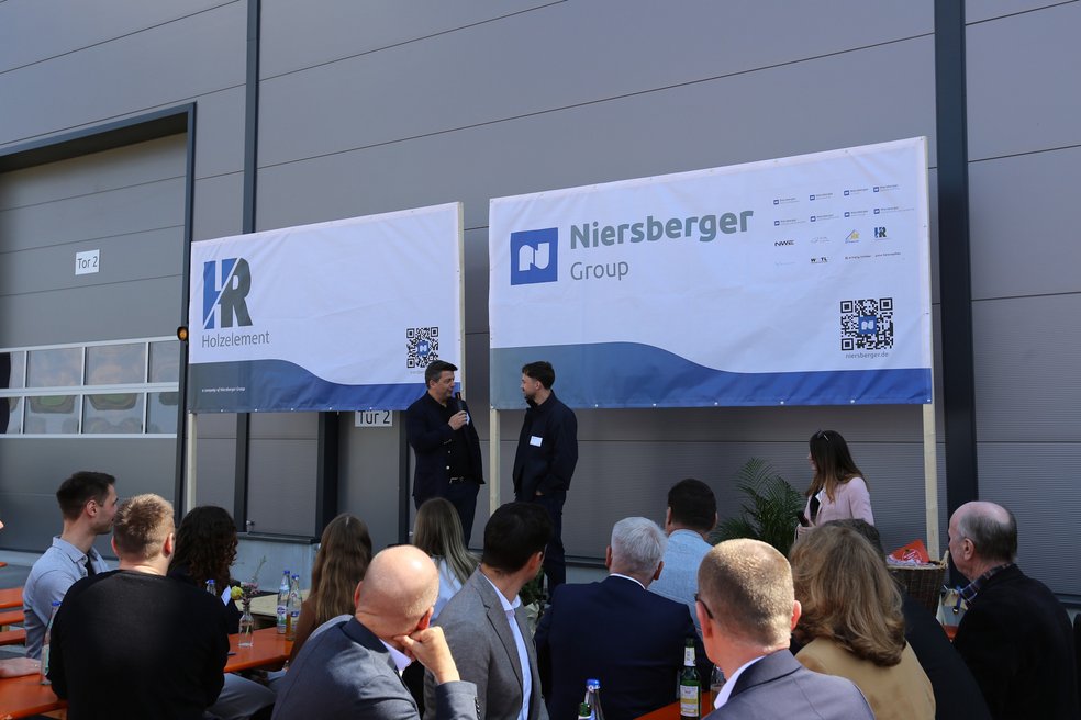 Zwei Personen im Gespräch auf einer Bühne vor einer Produktionshalle, davor sitzen Zuschauer an Biergartentischen.