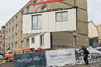 Fassadenelement schwebt vor einem Mehrfamilienhaus.