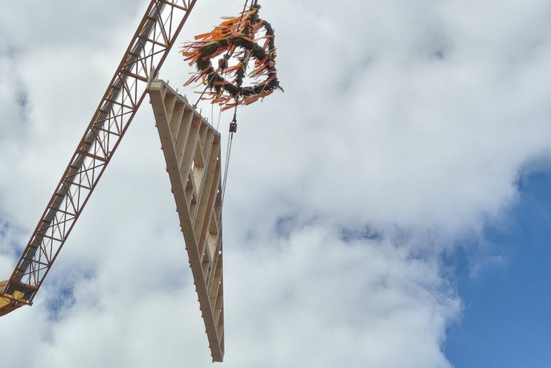 Dreieckiges vorgefertigtes Hausfassadenelement hängt an einem Kran, ebenso ein Richtkranz