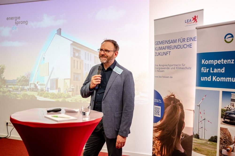 Person mit Mikrofon in der Hand steht an einem Stehtisch, im Hintergrund stehen Info-Banner.