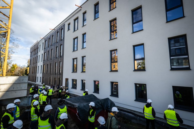 Personen mit gelben Warnwesten und weißen Bauhelmen stehen vor einem viergeschossigen Gebäude, dass gerade snaiert wird.